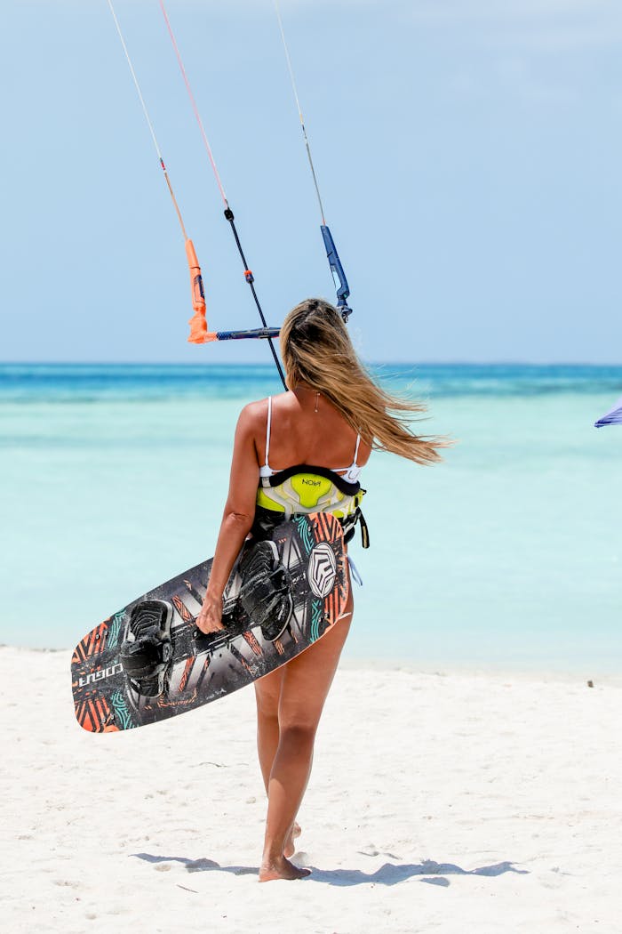A woman with long hair carries a kiteboard on a sunny beach.