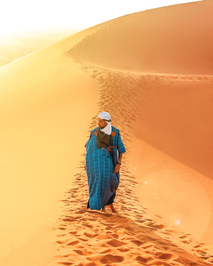 A person wearing traditional clothing walks across the sand dunes of the Sahara Desert at sunrise.